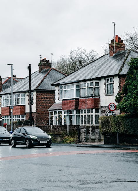 A residential street scene featuring two adjacent semi-detached houses with brick exteriors and pitched tiled roofs, situated behind a low fence and small front gardens. The house on the right has a w